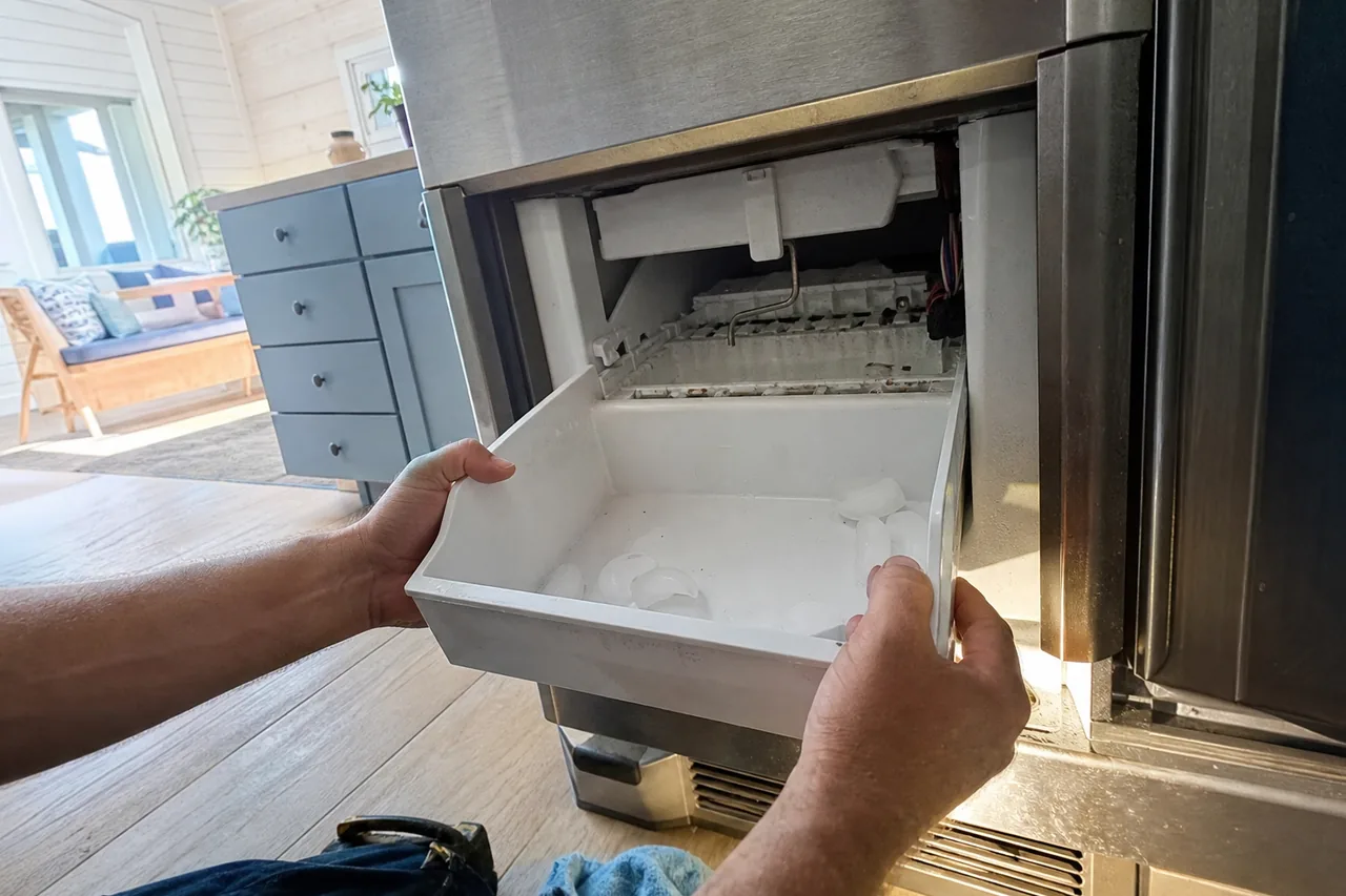 Two hands sliding a white plastic ice bin out from beneath a stainless ice maker, a few cubes left in the bin and the wire signal arm hanging down above the mold.